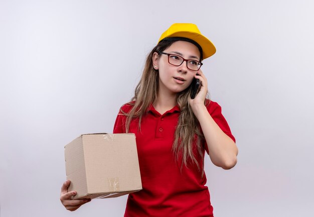 真面目な顔で携帯電話で話している間赤いポロシャツと黄色の帽子保持ボックスパッケージの若い配達の女の子