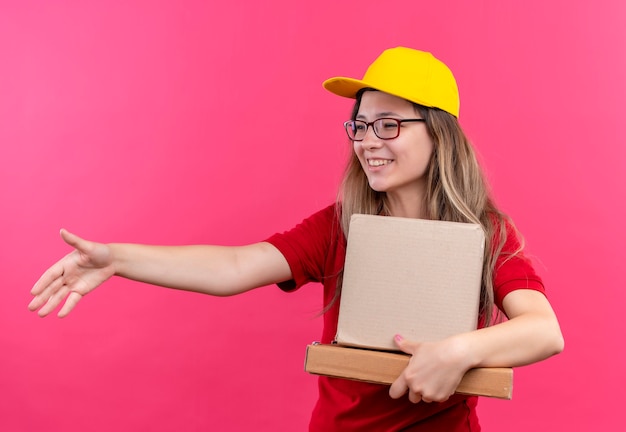 Молодая доставщица в красной рубашке поло и желтой кепке держит картонные коробки, приветствуя, предлагая улыбку руки