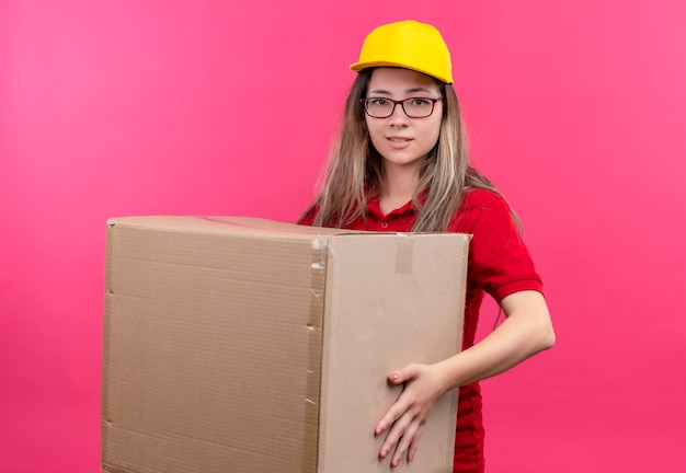 Young delivery girl in red polo shirt and yellow cap holding large cardboard box looking at camera smiling 