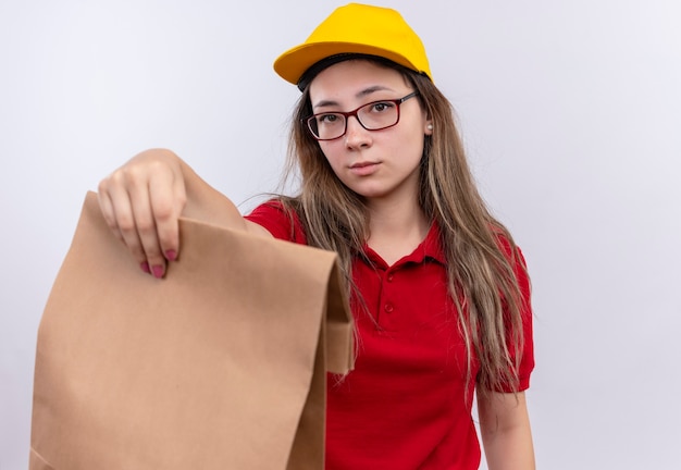 Foto gratuita giovane ragazza di consegna in camicia di polo rossa e cappuccio giallo che tiene il pacchetto di carta che guarda l'obbiettivo con l'espressione sicura seria