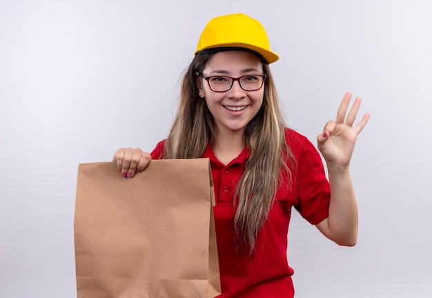 赤いポロシャツと黄色い帽子の若い配達の女の子が元気にOKサインを示して笑っている紙のパッケージを保持