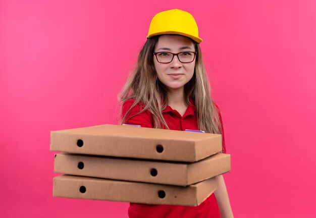 Young delivery girl in red polo shirt and yellow cap holding stack of pizza boxes looking at camera with confident smile on face 