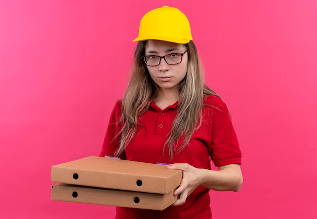 Young delivery girl in red polo shirt and yellow cap holding stack of pizza boxes looking at camera with sad expression on face 