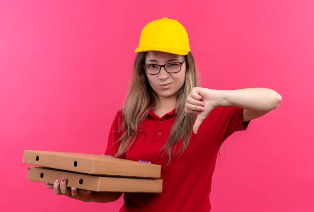 Young delivery girl in red polo shirt and yellow cap holding stack of pizza boxes showing dislike 