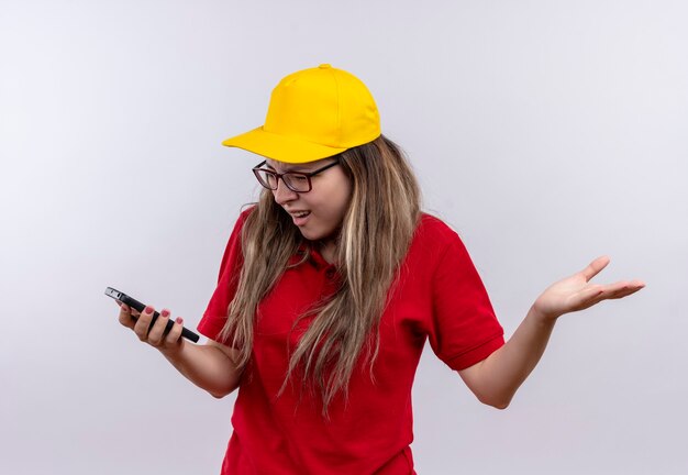 Young delivery girl in red polo shirt and yellow cap looking at screen of her smartphone frustrated 