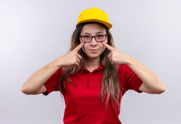 Young delivery girl in red polo shirt and yellow cap pointing with index fingers to her eyes smiling 