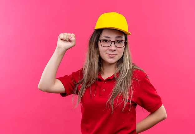 Free photo young delivery girl in red polo shirt and yellow cap positive and happy clenching fist, winner concept