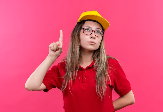 Young delivery girl in red polo shirt and yellow cap showing index finger  having  new idea 