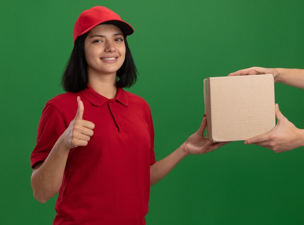 Young delivery girl in red uniform and cap giving cardboard box to a customer smiling friendly showing thumbs up standing over green wall