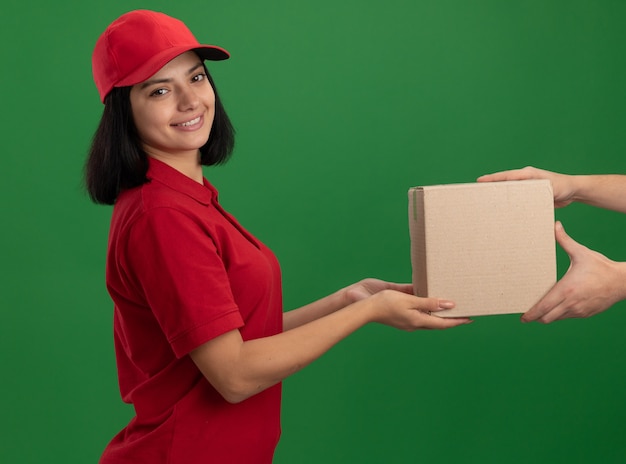 緑の壁の上に立ってフレンドリーな笑顔の顧客に段ボール箱を与える赤い制服とキャップの若い配達の女の子