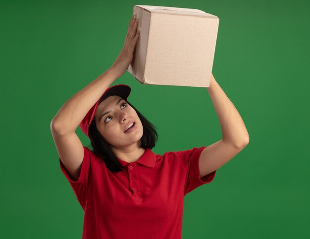 Free photo young delivery girl in red uniform and cap holding cardboard box over her head looking at it with interest standing over green wall