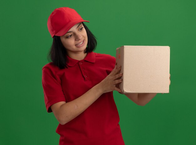 빨간 제복을 입은 젊은 배달 소녀와 골판지 상자를 들고 모자는 녹색 벽 위에 서서 흥미를 느낍니다.