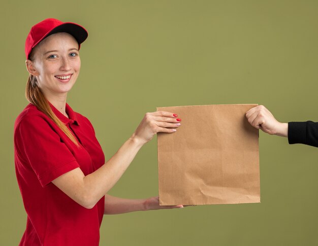 Молодая доставщица в красной форме и кепке держит бумажный пакет, дарит его клиенту, уверенно улыбаясь над зеленой стеной