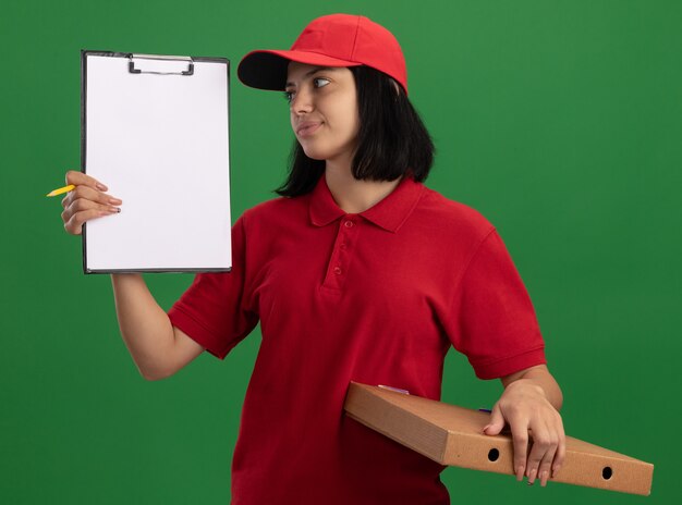 Молодая доставщица в красной форме и кепке держит коробку для пиццы и буфер обмена с карандашом, глядя на нее с улыбкой на лице, стоя над зеленой стеной