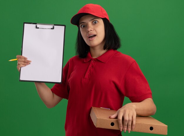 Молодая доставщица в красной форме и кепке держит коробку для пиццы, показывая буфер обмена с удивленным карандашом, стоя над зеленой стеной