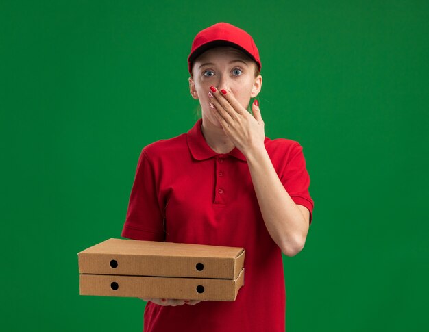 Young delivery girl in red uniform and cap holding pizza boxes  being shocked covering mouth with hand standing over green wall