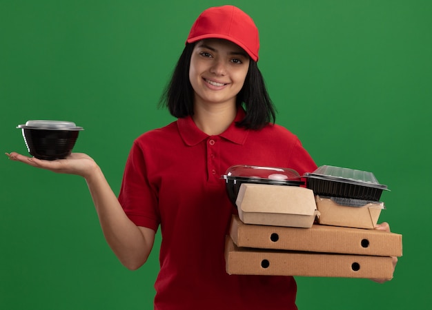 Foto gratuita giovane ragazza di consegna in uniforme rossa e cappuccio che tiene scatole per pizza e confezioni di cibo con un sorriso sul viso in piedi sopra la parete verde