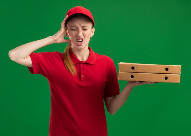 빨간색 유니폼과 모자를 들고 젊은 배달 소녀 녹색 벽 위에 서 실수로 그녀의 머리에 손으로 혼란 찾고 피자 상자를 들고