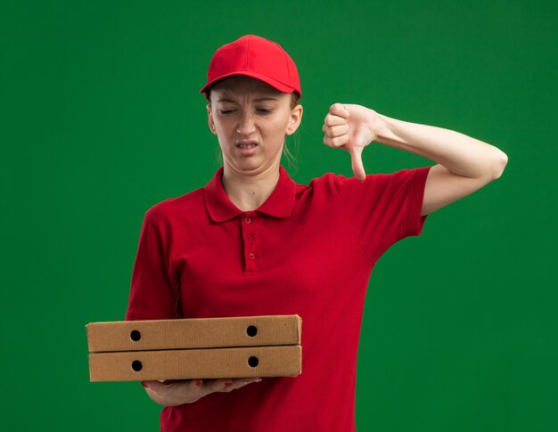 Young delivery girl in red uniform and cap holding pizza boxes looking at them displeased showing thumbs down standing over green wall