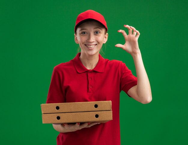 빨간 제복을 입은 젊은 배달 소녀와 모자는 녹색 벽 위에 유쾌하게 서있는 손가락으로 작은 크기의 제스처를 만드는 피자 상자를 들고