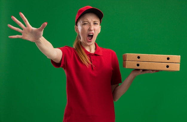 Молодая доставщица в красной униформе и кепке держит коробки для пиццы и кричит с агрессивным выражением лица, делая жест остановки рукой, стоящей над зеленой стеной