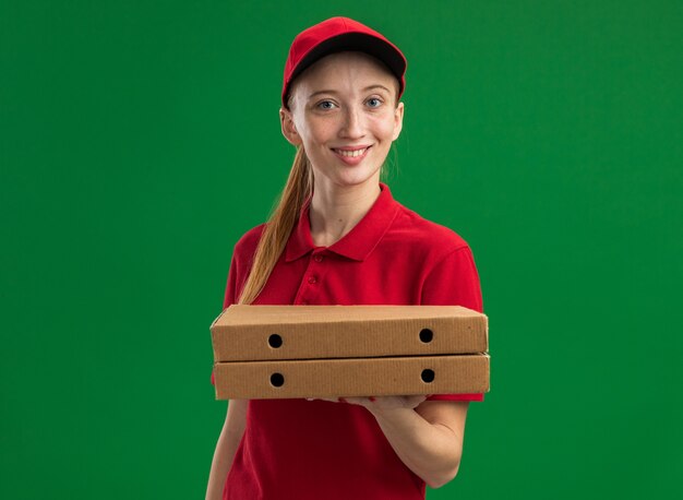 Молодая доставщица в красной форме и кепке держит коробки для пиццы с улыбкой на счастливом лице, стоящем над зеленой стеной