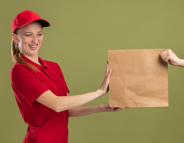 Young delivery girl in red uniform and cap refusing to receive paper package standing over green wall