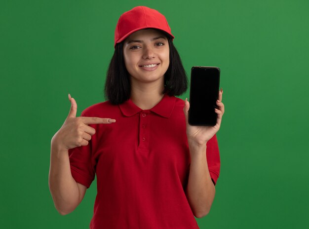 Young delivery girl in red uniform and cap showing smartphone pointing with index finger at it smiling confident standing over green wall