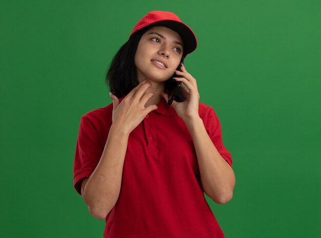 Young delivery girl in red uniform and cap smiling while talking on mobile phone standing over green wall