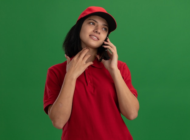 Молодая доставщица в красной форме и кепке улыбается во время разговора по мобильному телефону, стоя над зеленой стеной