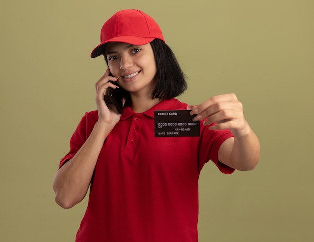 赤い制服を着た若い配達の女の子と携帯電話で話しているキャップは、明るい壁の上に元気に立って笑顔のクレジットカードを示しています