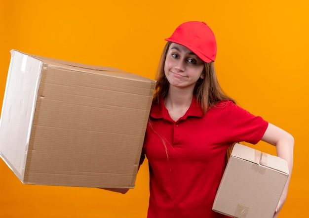 Young delivery girl in red uniform holding boxes on isolated orange wall