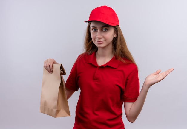 Giovane ragazza di consegna in uniforme rossa che tiene il sacchetto di carta e che mostra la mano vuota sulla parete bianca isolata