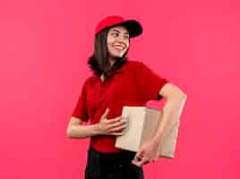 Free photo young delivery girl wearing red polo shirt and cap holding box package looking aside with happy smile on face standing over pink background
