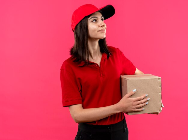 Young delivery girl wearing red polo shirt and cap holding cardboard box package looking aside with smile on face standing over pink wall
