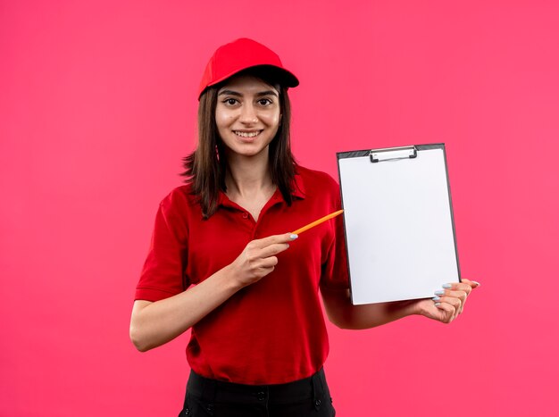 ピンクの背景の上に立っている幸せそうな顔で笑ってそれを鉛筆で指している空白のページでクリップボードを保持している赤いポロシャツとキャップを身に着けている若い配達の女の子