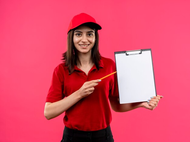 Young delivery girl wearing red polo shirt and cap holding clipboard with blank pages pointing with pencil at it smiling with happy face standing over pink background