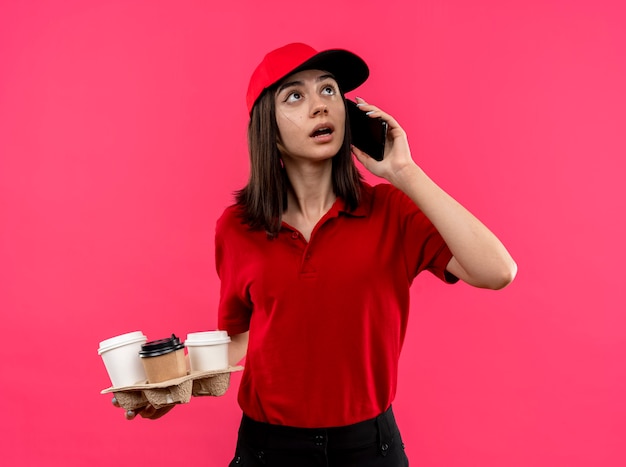 Giovane ragazza delle consegne che indossa la maglietta polo rossa e cappuccio che tiene il pacchetto alimentare che sembra confuso mentre parla al telefono cellulare in piedi sopra il muro rosa