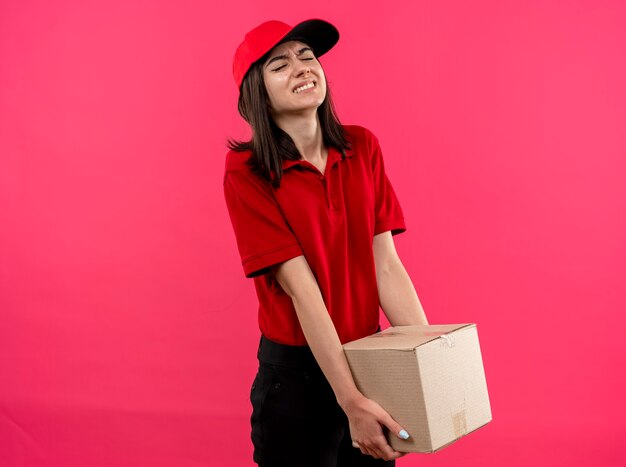 Молодая доставщица в красной рубашке поло и кепке с тяжелой коробкой выглядит усталой и напряженной, стоя у розовой стены