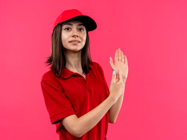 Young delivery girl wearing red polo shirt and cap looking at camera smiling pointing at her arm with finger standing over pink background