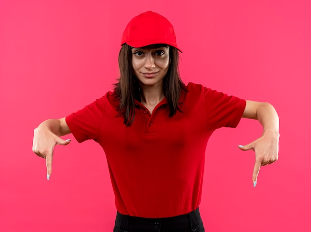 Free photo young delivery girl wearing red polo shirt and cap pointing wityh index fingers down smiling confident standing over pink wall