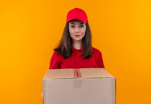 Молодая доставщица в красной футболке в красной кепке держит большую коробку на изолированном желтом фоне