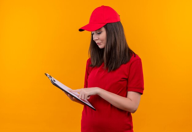격리 된 노란색 배경에 클립 보드를 들고 빨간 모자에 빨간 티셔츠를 입고 젊은 배달 소녀