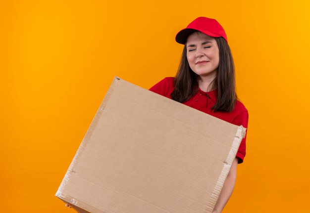 Young delivery girl wearing red t-shirt in red cap holding heavy box on isolated yellow background