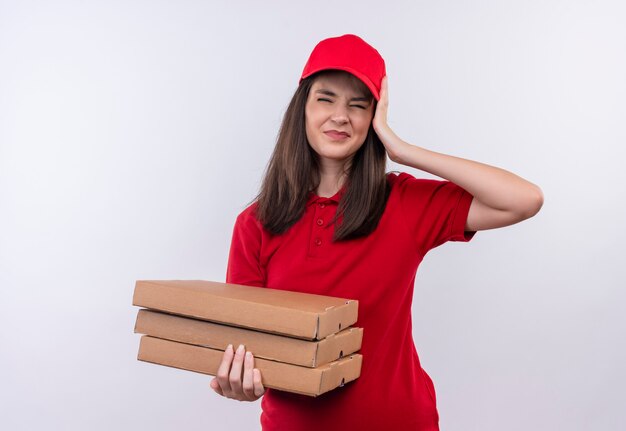 Молодая доставщица в красной футболке в красной кепке держит коробку для пиццы и схватила головы на изолированном белом фоне