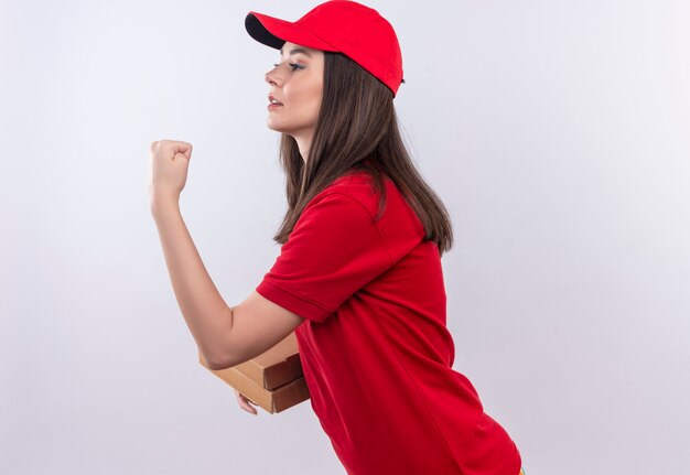 Молодая доставщица в красной футболке в красной кепке держит коробку для пиццы и показывает кулак на изолированном белом фоне