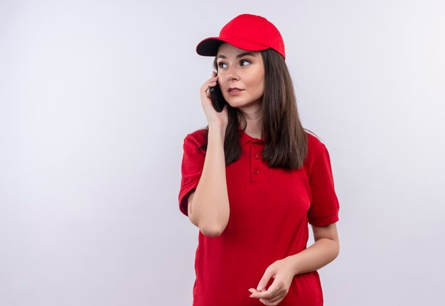 Молодая доставщица в красной футболке в красной кепке делает телефонный звонок на изолированном белом фоне