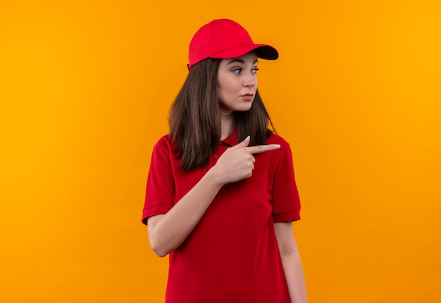 Young delivery girl wearing red t-shirt in red cap points a finger to the side on isolated yellow background