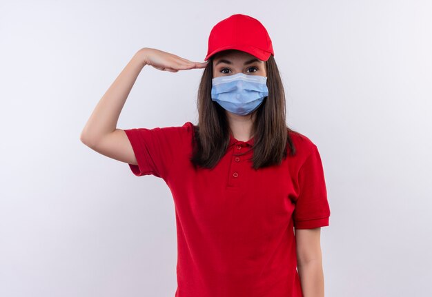 Young delivery girl wearing red t-shirt in red cap wears face mask and give salute on isolated white background