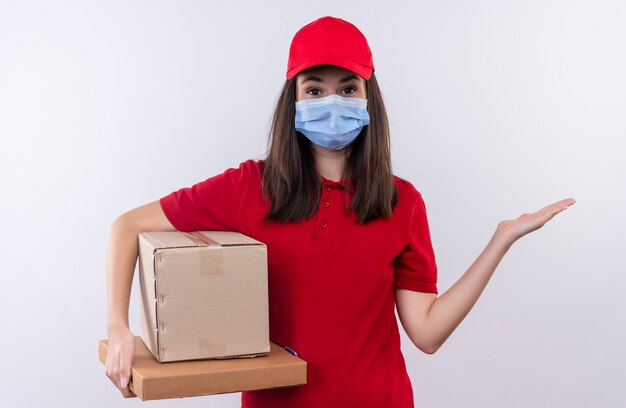 La giovane ragazza di consegna che porta la maglietta rossa nel cappuccio rosso indossa la maschera facciale che tiene una scatola e una scatola della pizza su fondo bianco isolato